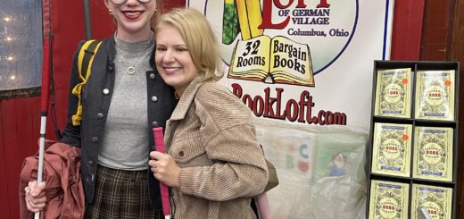 Cass & Case stand smiling in front of a sign for 'The Book Loft of German Village' in Columbus, Ohio. Both are holding white canes and stand among colorful signage and stacks of books wrapped in plastic.