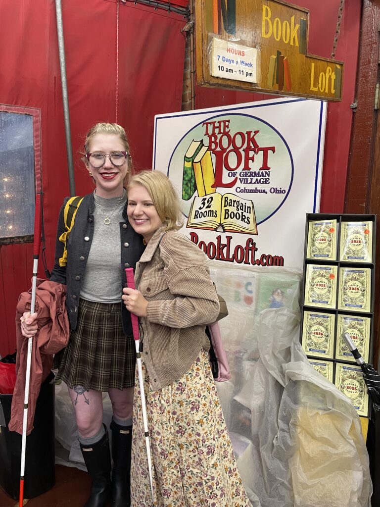 Cass & Case stand smiling in front of a sign for 'The Book Loft of German Village' in Columbus, Ohio. Both are holding white canes and stand among colorful signage and stacks of books wrapped in plastic.