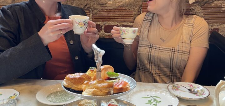 Cass (left) and Case (right) enjoy an afternoon tea. They smile warmly and toast with delicate teacups in front of a tiered tray of sandwiches and pastries.