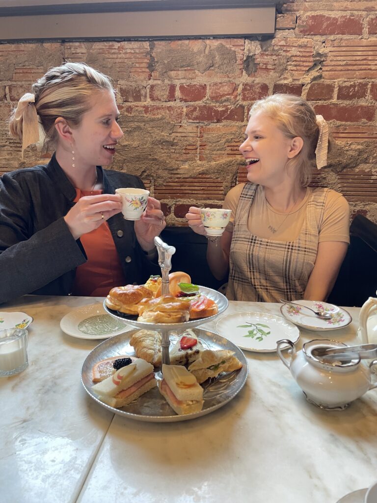 Cass (left) and Case (right) enjoy an afternoon tea. They smile warmly and toast with delicate teacups in front of a tiered tray of sandwiches and pastries.