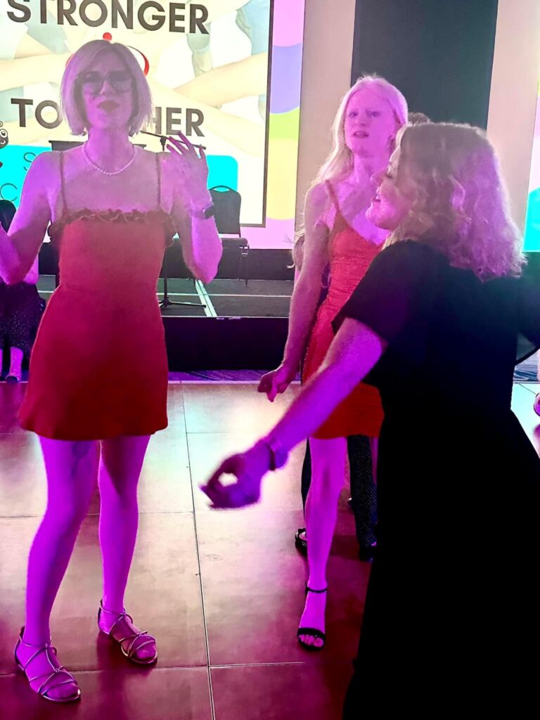 Case, Cass, and a friend of theirs dancing on a lit dance floor, illuminated by colorful lighting that gives the scene a vibrant, purple-pink hue. The background features a stage with chairs and a large digital screen displaying the words "STRONGER TOGETHER" in bold letters.