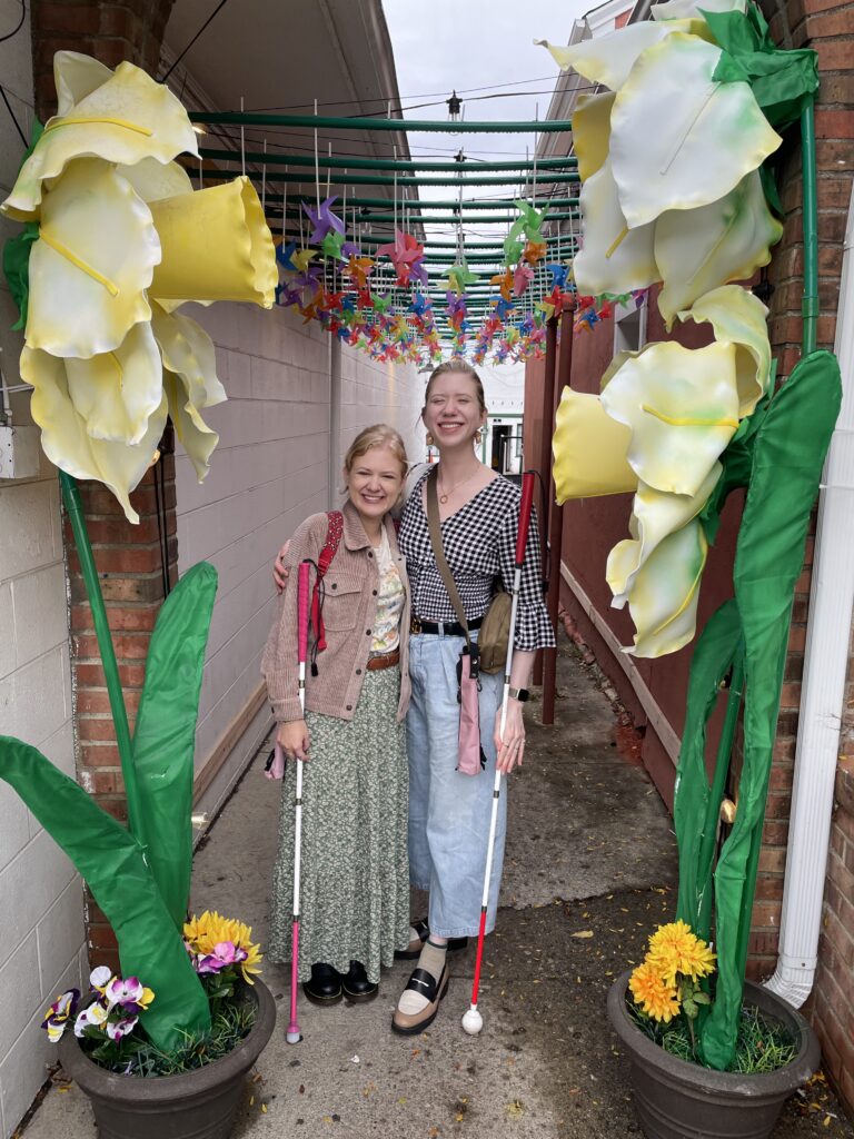 case and cass smile and stand together in a colorful outdoor walkway decorated with large artificial yellow and white flowers and hanging multicolored pinwheels. both women hold their white canes and stand closely with their arms around each other