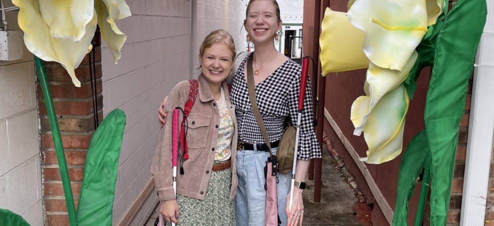 case and cass smile and stand together in a colorful outdoor walkway decorated with large artificial yellow and white flowers and hanging multicolored pinwheels. both women hold their white canes and stand closely with their arms around each other