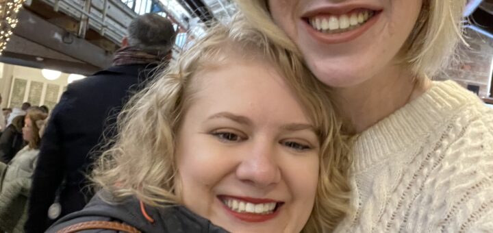 selfie of case and cass at Chelsea Market in New York. they smile widely, their heads close together in front of a lit archway made of string lights