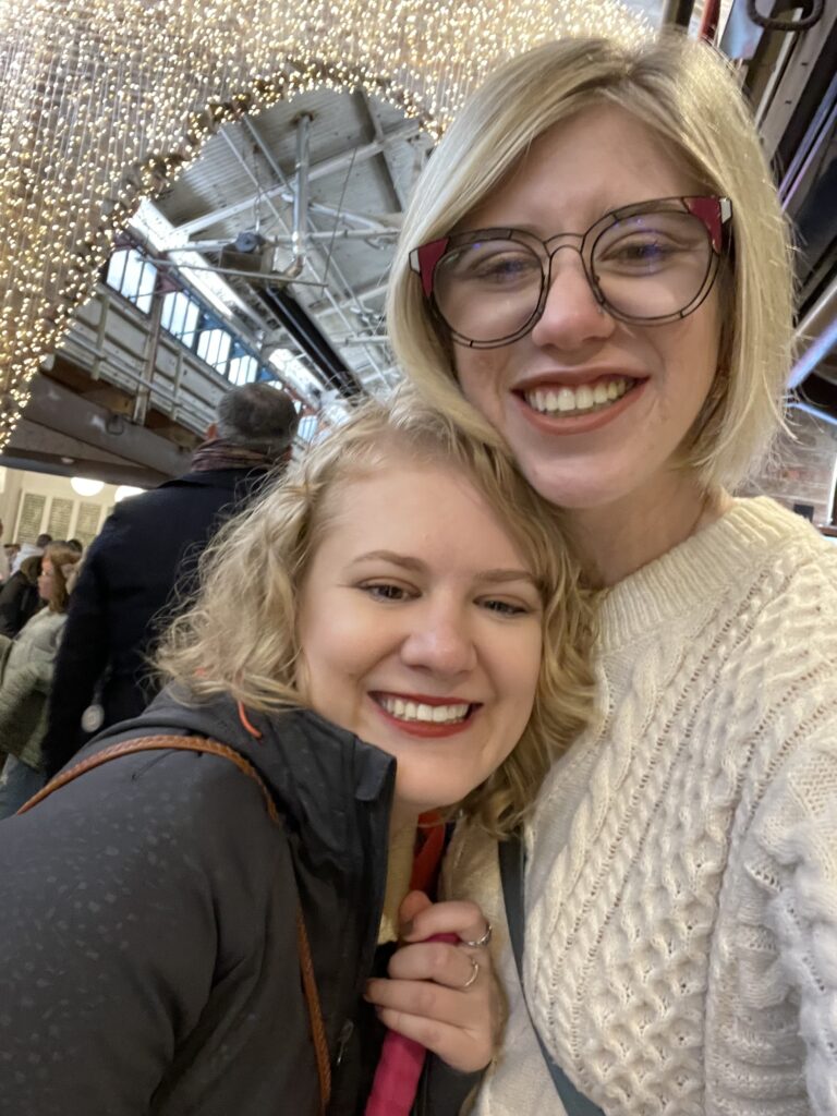 selfie of case and cass at Chelsea Market in New York. they smile widely, their heads close together in front of a lit archway made of string lights