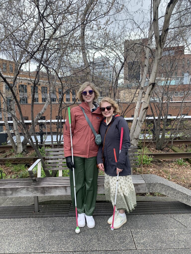 Cass (left) and Case (right) stand together outdoors on a paved walkway, smiling at the camera. Both are holding white canes. Behind them are leafless trees, a wooden bench, and a cityscape with brick buildings under an overcast sky