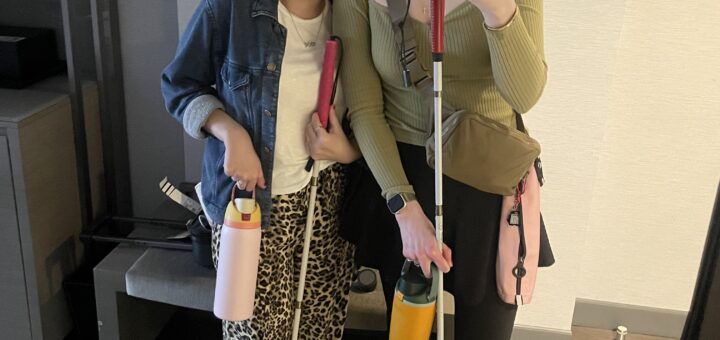 Case + Cass stand in front of a mirror in their hotel room, taking a selfie. Both are holding white canes and water bottles.