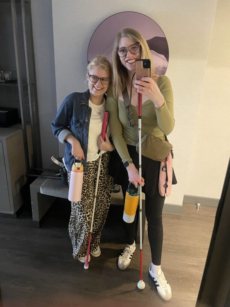 Case + Cass stand in front of a mirror in their hotel room, taking a selfie. Both are holding white canes and water bottles.