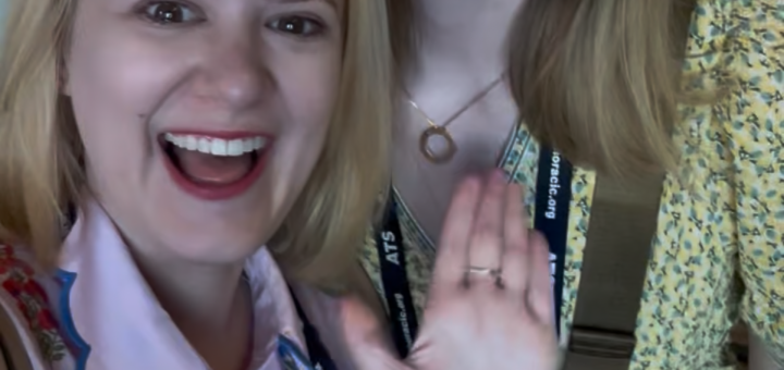 Case + Cass stand close together indoors, smiling and waving at the camera