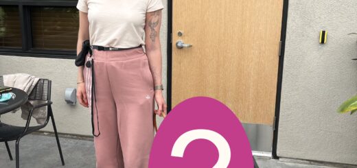 Cass stands outside on a patio. She is smiling, with one hand holding a leather leash connected to something obscured by a large pink oval with a white question mark covering the lower part of the image.