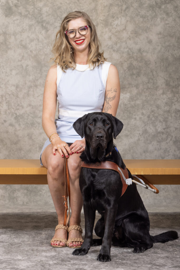 Cass, with wavy blonde hair, red lipstick, and glasses sits on a wooden bench wearing a light blue sleeveless dress and tan braided sandals. Beside her sits a tall, black Labrador retriever wearing a brown guide dog harness. The two pose in front of a neutral gray backdrop.