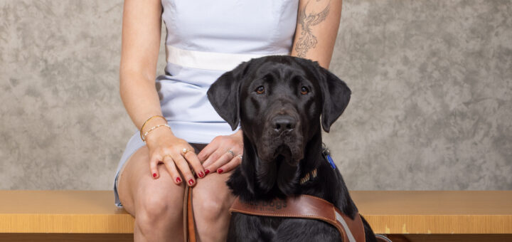 Cass, with wavy blonde hair, red lipstick, and glasses sits on a wooden bench wearing a light blue sleeveless dress and tan braided sandals. Beside her sits a tall, black Labrador retriever wearing a brown guide dog harness. The two pose in front of a neutral gray backdrop.