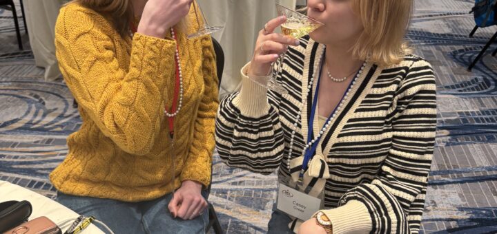 Case and Cass sit at a round table in a conference room, each sipping sparkling cider from a martini glass and smiling