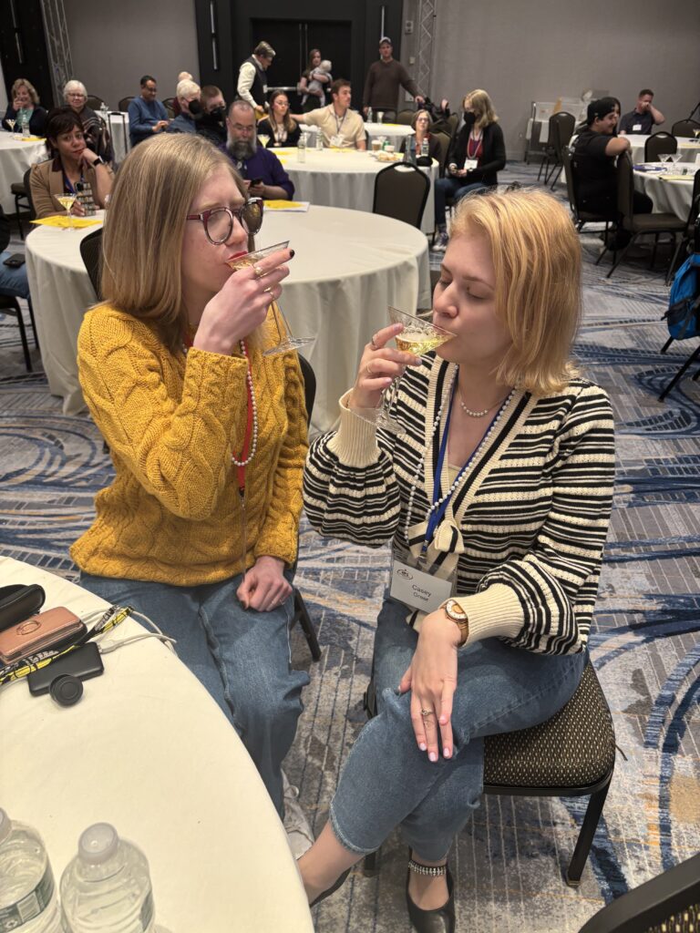 Case and Cass sit at a round table in a conference room, each sipping sparkling cider from a martini glass and smiling