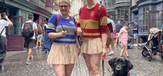 Case & Cass stand in Diagon Alley at Universal Studios, each holding a wand. They wear matching beige skirts and striped Hogwarts house sweaters—Case in blue and grey, Cass in red and gold. Case holds her pink-handled cane and a black lab guide dog sits at the right of Cass' feet, looking toward the camera. Behind them, detailed wizarding shop facades line the street, with a large dragon perched atop Gringotts Bank in the background.