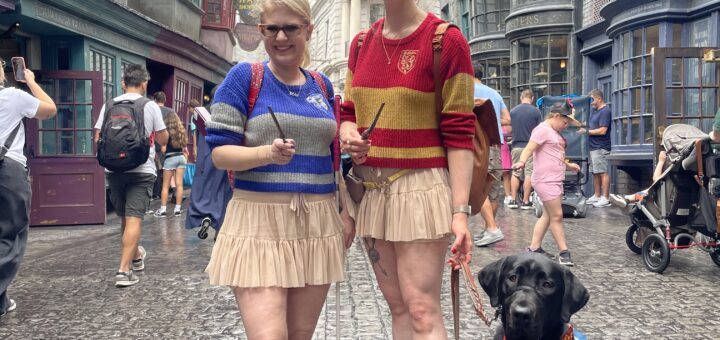 Case & Cass stand in Diagon Alley at Universal Studios, each holding a wand. They wear matching beige skirts and striped Hogwarts house sweaters—Case in blue and grey, Cass in red and gold. Case holds her pink-handled cane and a black lab guide dog sits at the right of Cass' feet, looking toward the camera. Behind them, detailed wizarding shop facades line the street, with a large dragon perched atop Gringotts Bank in the background.