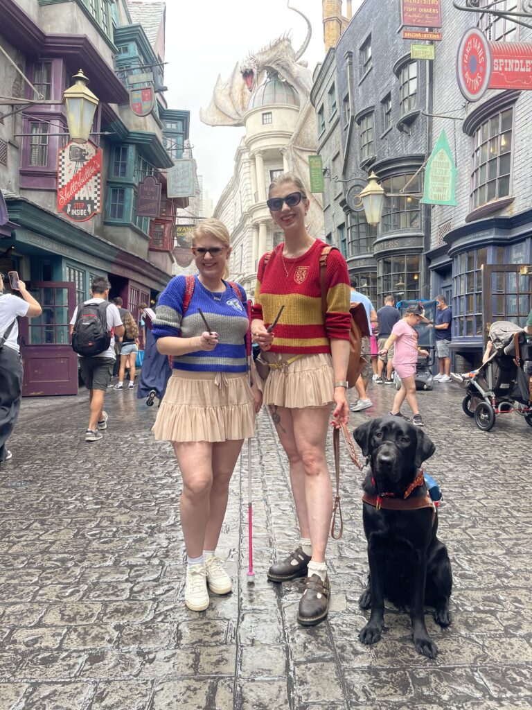 Case & Cass stand in Diagon Alley at Universal Studios, each holding a wand. They wear matching beige skirts and striped Hogwarts house sweaters—Case in blue and grey, Cass in red and gold. Case holds her pink-handled cane and a black lab guide dog sits at the right of Cass' feet, looking toward the camera. Behind them, detailed wizarding shop facades line the street, with a large dragon perched atop Gringotts Bank in the background.