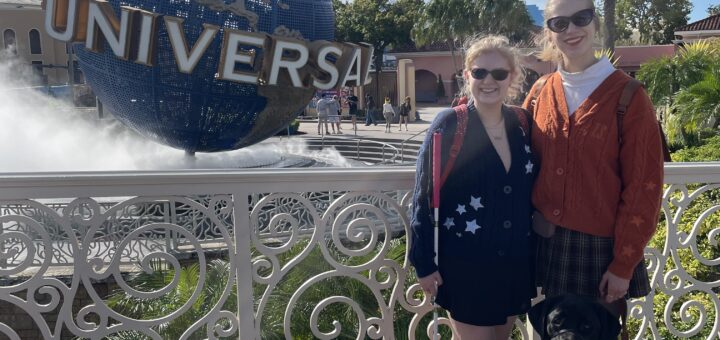 Case and Cass stand smiling in front of the Universal Studios globe fountain on a sunny day. Case holds a white cane, and a black guide dog sits beside Cass.