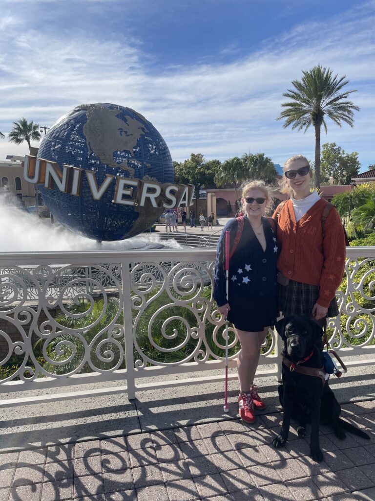 Case and Cass stand smiling in front of the Universal Studios globe fountain on a sunny day. Case holds a white cane, and a black guide dog sits beside Cass.