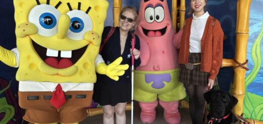 Case and Cass stand smiling between costumed SpongeBob SquarePants and Patrick Star at a theme park, with jellyfish decor overhead. Case holds a white cane, and a black guide dog sits at Cass’s feet.
