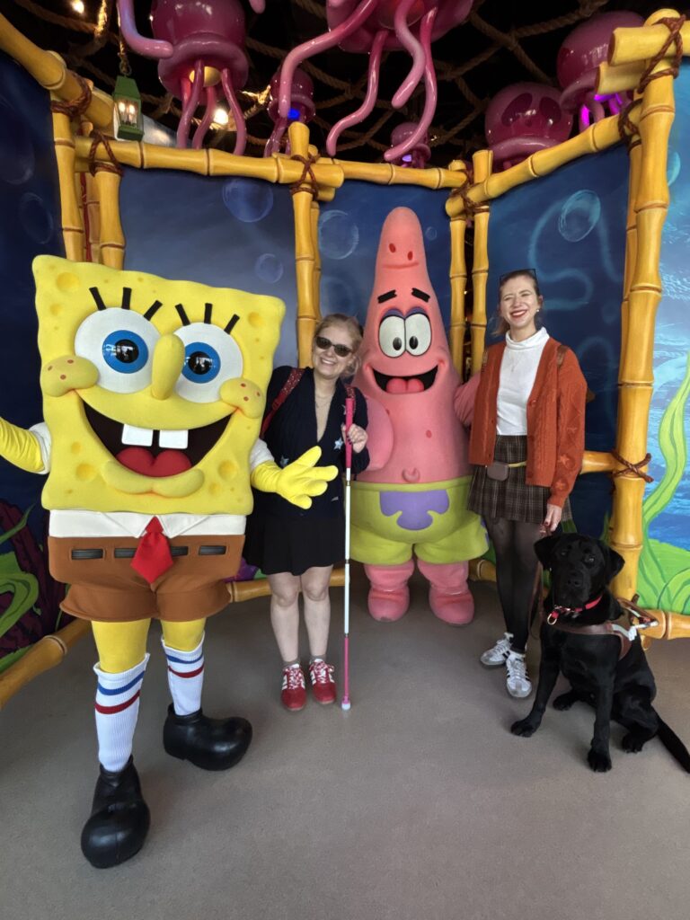 Case and Cass stand smiling between costumed SpongeBob SquarePants and Patrick Star at a theme park, with jellyfish decor overhead. Case holds a white cane, and a black guide dog sits at Cass’s feet.
