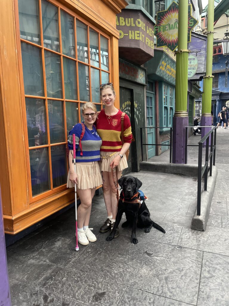Two best friends stand smiling together on a colorful, whimsical street with bright storefronts in orange, teal, and purple. Both wear matching striped sweaters and pleated skirts. The friend on the left holds a white cane with a pink tip, and the friend on the right stands beside a black guide dog wearing a harness. The dog sits calmly at their feet as they pose closely with their arms around each other.