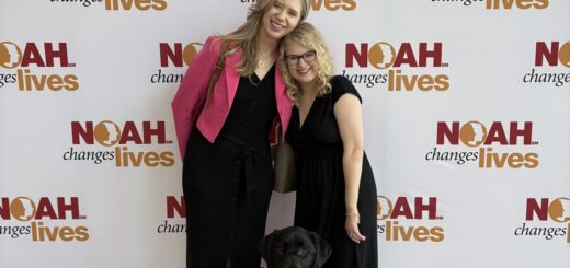 Cassandra and Casey smile together at the 2026 albinism conference with Cassandra's black guide dog sitting in front of them against a NOAH event backdrop