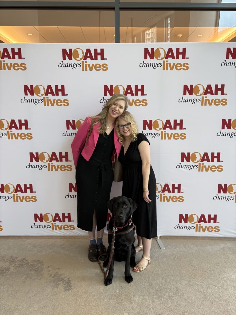 Cassandra and Casey smile together at the 2026 albinism conference with Cassandra's black guide dog sitting in front of them against a NOAH event backdrop