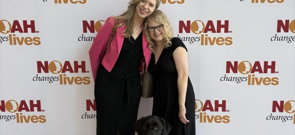 Cassandra and Casey smile together at the 2026 albinism conference with Cassandra's black guide dog sitting in front of them against a NOAH event backdrop