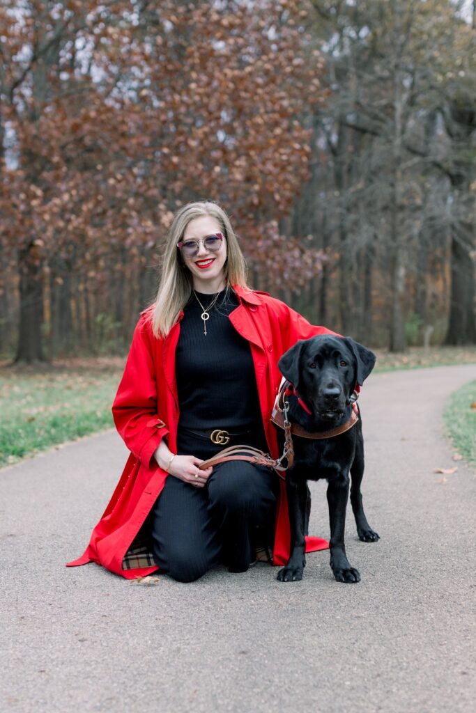 photo of Cassandra, a smiling tall woman with fair skin and shoulder-length blonde hair. Wearing a black outfit and long red coat, she kneels with her arm around her tall black lab guide dog Anaheim standing beside her