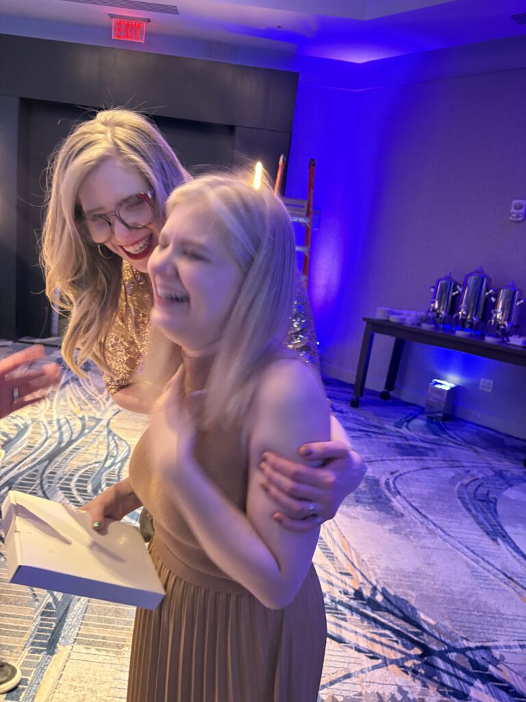 in a slightly blurry photo, Case and Cass laugh and embrace at a conference, dressed up for the evening.