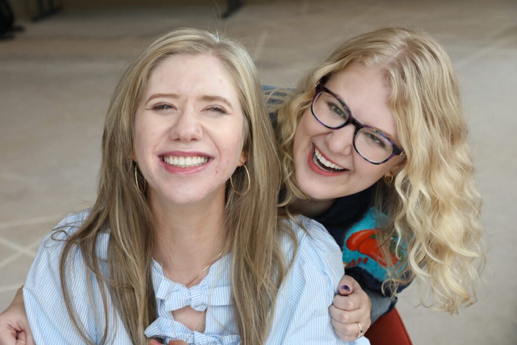 Cass and Case smile and laugh together for the camera. Cass wears a blue-and-white striped shirt with a bow, while Case wears glasses and a colorful shirt.
