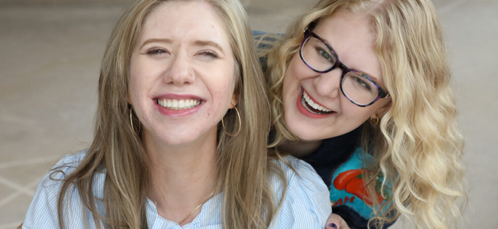 Cass and Case smile and laugh together for the camera. Cass wears a blue-and-white striped shirt with a bow, while Case wears glasses and a colorful shirt.