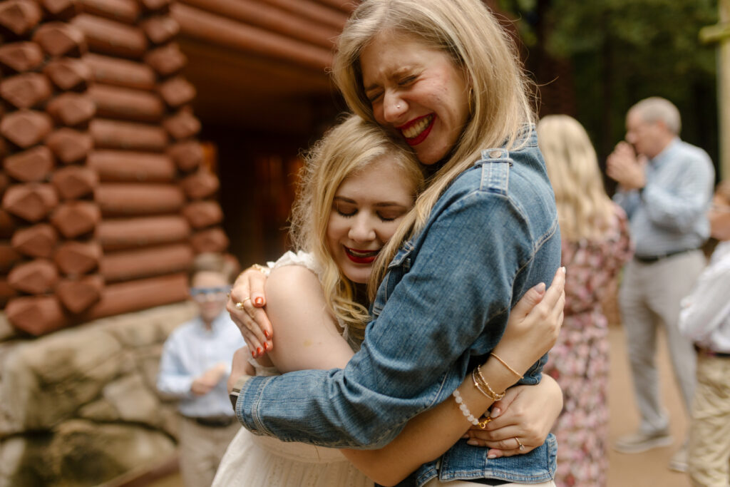 Case, in a white dress, and Cass, in a denim jacket, embrace with their eyes closed and big smiles outside a log cabin wall