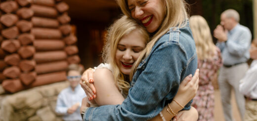 Case, in a white dress, and Cass, in a denim jacket, embrace with their eyes closed and big smiles outside a log cabin wall