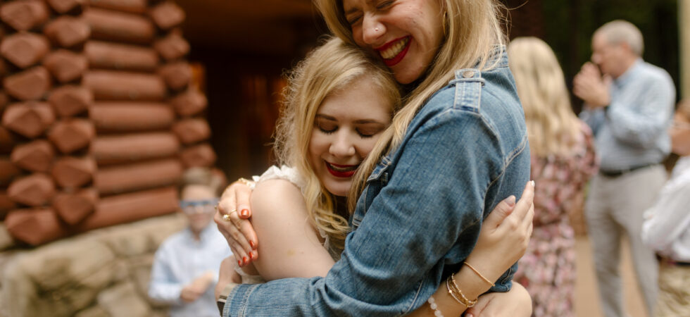 Case, in a white dress, and Cass, in a denim jacket, embrace with their eyes closed and big smiles outside a log cabin wall